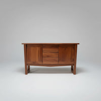 A mid-century modern wooden sideboard or credenza with a warm reddish-brown finish, featuring a central drawer section and angled legs, photographed in a cluttered warehouse or furniture workshop setting.