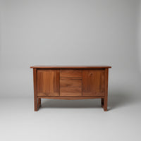 A mid-century modern wooden sideboard or credenza with three drawers, shown in a cluttered warehouse or furniture showroom setting.