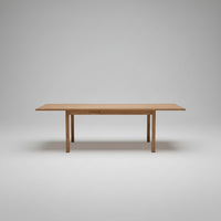 A rectangular wooden dining table with a live-edge design and a visible center seam, shown in a modern interior setting with a concrete floor and staircase in the background.