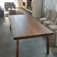 A rectangular wooden dining table with a medium brown finish and visible wood grain, shown in a modern living room setting with chairs and a sofa in the background.