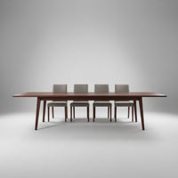 A long rectangular wooden conference table with a dark reddish-brown finish and slim legs, shown in a modern room with sheer curtains, four chairs, and a concrete floor.