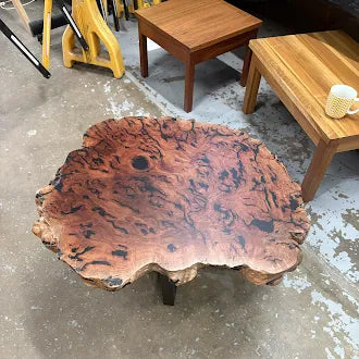 A rustic live-edge coffee table made from a richly patterned burl wood slab, displayed on a concrete floor in a furniture showroom.