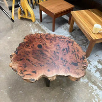 A rustic live-edge coffee table made from a richly patterned burl wood slab, displayed on a concrete floor in a furniture showroom.