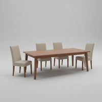 A long rectangular wooden dining table with a medium brown finish, shown in a room with a concrete floor and neutral upholstered chairs against sheer curtains.