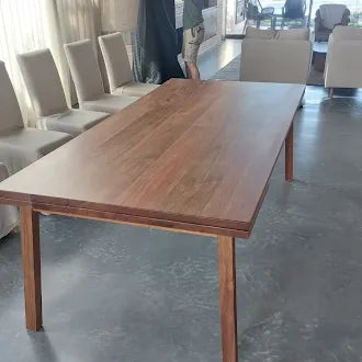 A rectangular wooden dining table with a warm medium-brown finish and visible wood grain, shown in a showroom or dining room setting with chairs and a gray floor.