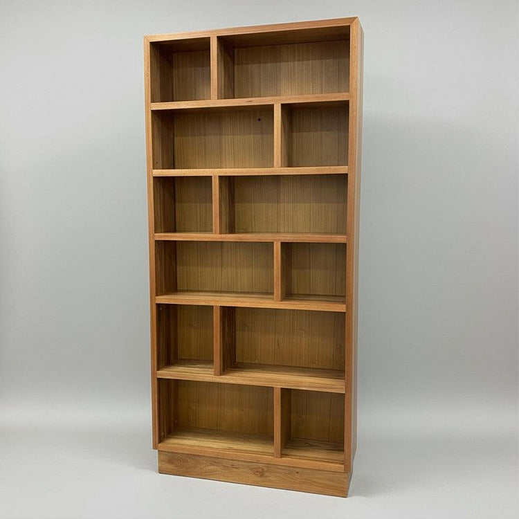 An asymmetrical wooden bookcase with multiple shelves, featuring a stool in front of it and a larger bookcase in the background.